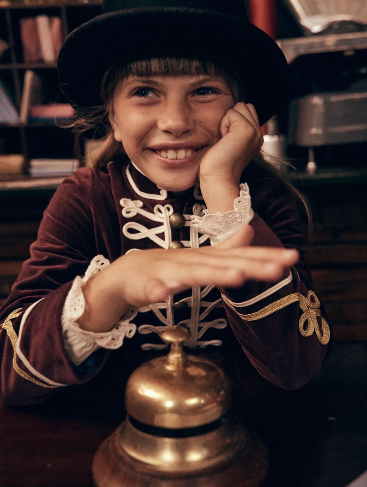 Girl in a velvet military-style jacket and top hat ringing a brass desk bell, Mini Boden Polar Post Winter 2026, styled by Charlotte Kewley
