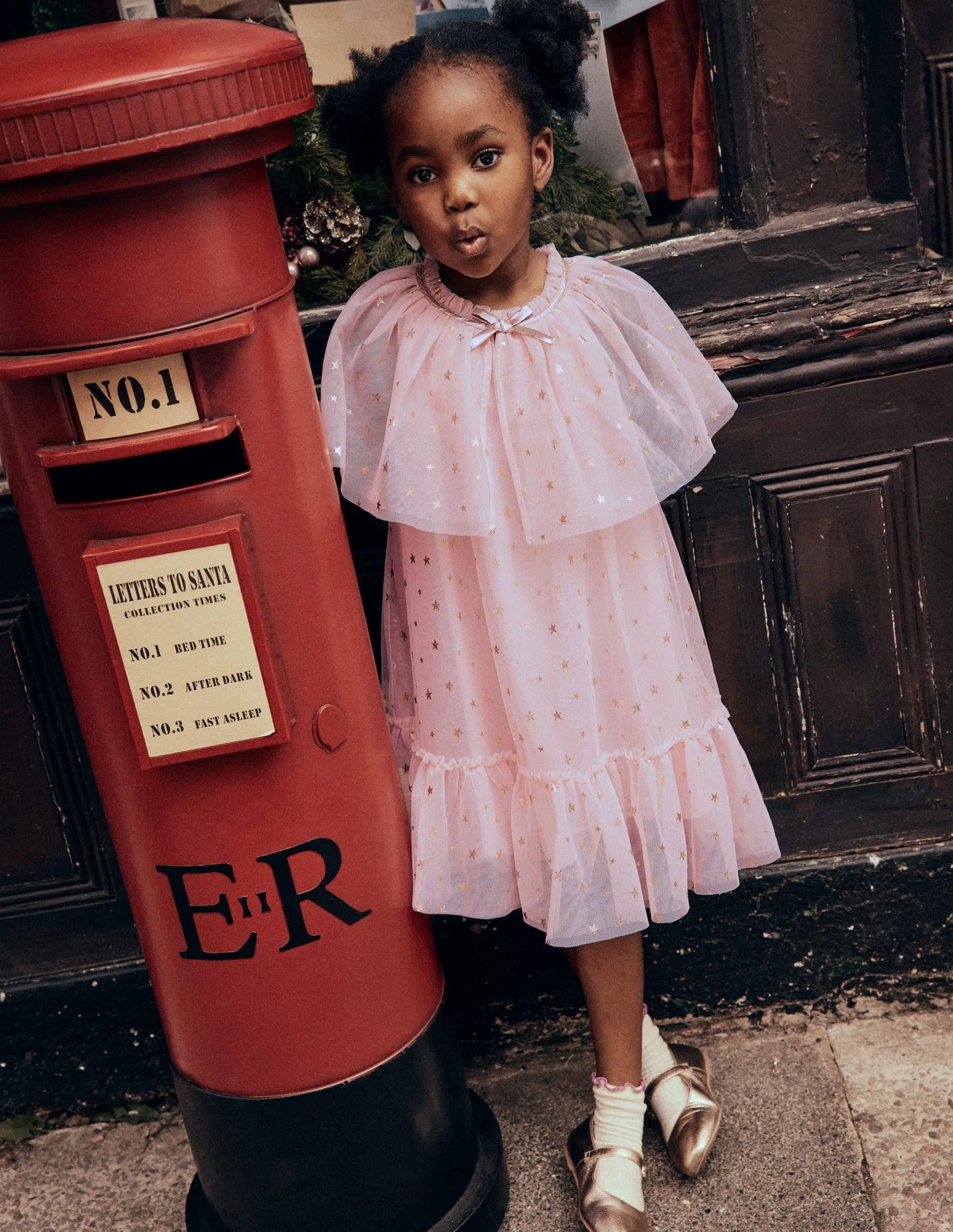 Girl in a pale pink tulle cape dress beside a red Letters to Santa post box, Mini Boden Polar Post Winter 2026