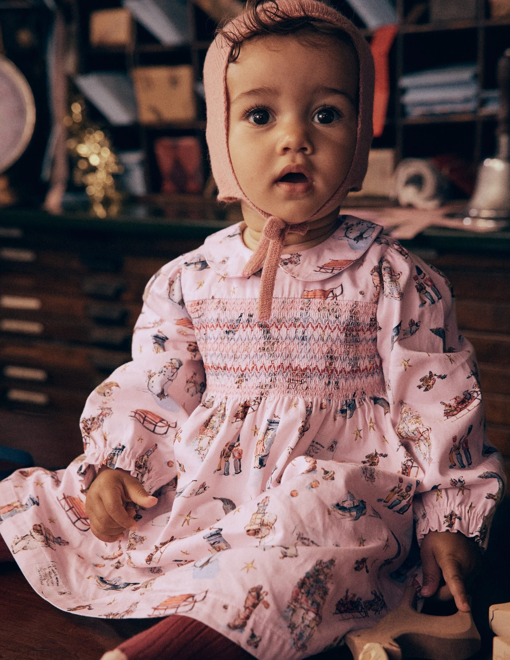 Baby in a pink smocked print dress and bonnet, Mini Boden Polar Post Winter 2026