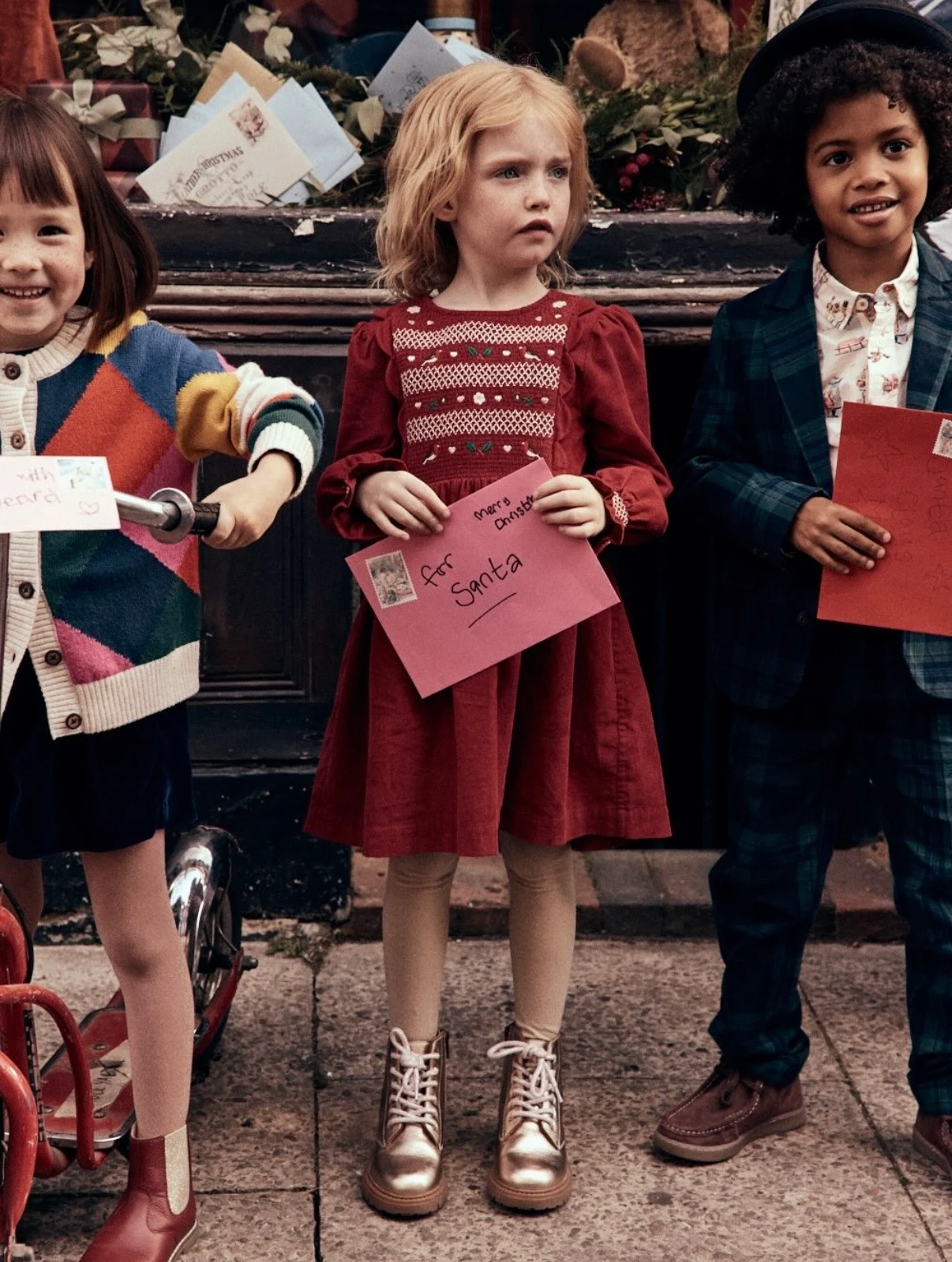 Three children holding letters to Santa outside the post office, Mini Boden Polar Post Winter 2026